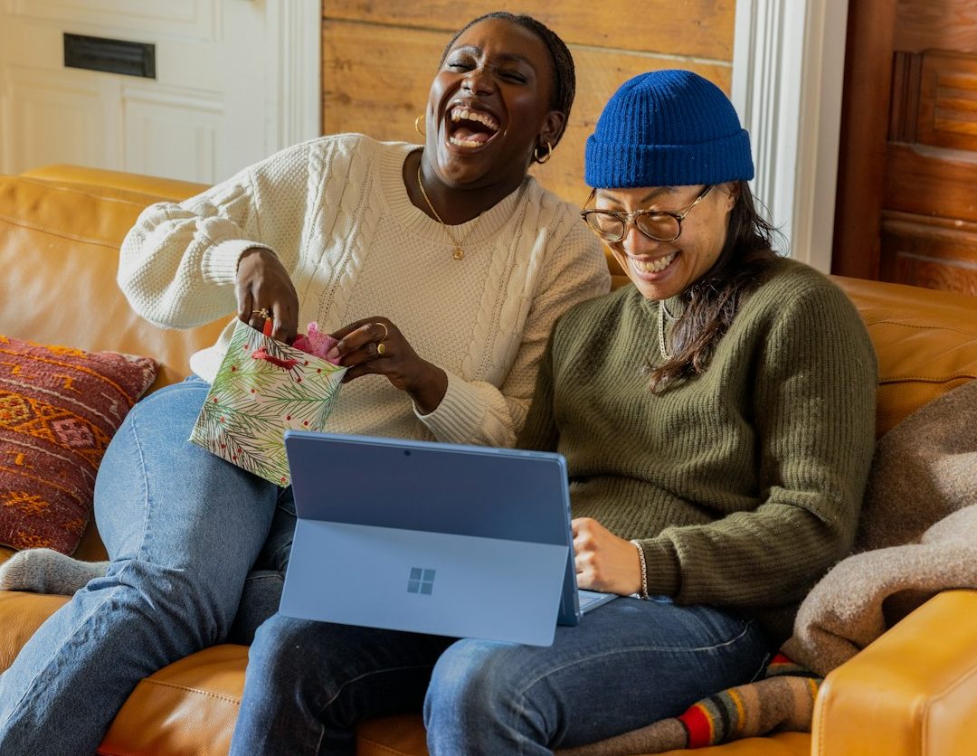 Wer zuletzt lacht, schreibt bessere Texte als KI! Zwei Frauen sitzen lachend auf einer Couch und schauen auf einen Laptop.