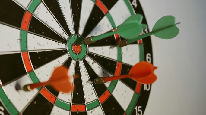 Dartboard mit Pfeilen in verschiedenen Farben, einige stecken im Ziel.